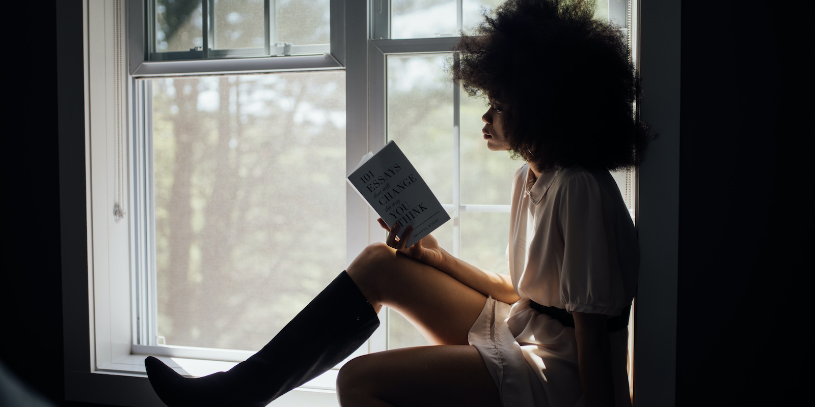 An young black woman with an afro sits on a shady window sill reading a book