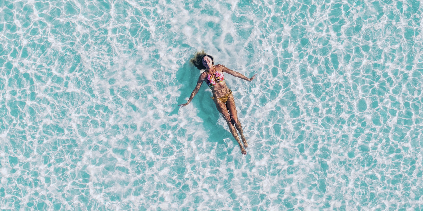An overhead photograph of a woman in a bikini floating on a foamy blue surface.