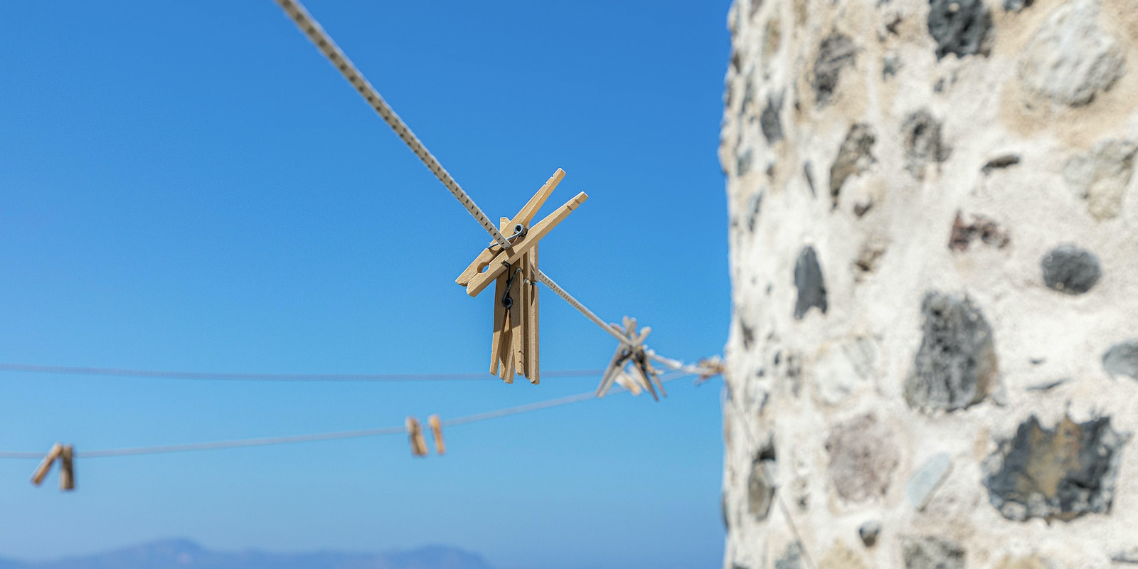 A photograph of an empty clothesline with wooden clothespins next to a tall stone wall in front of a clear blue sky.