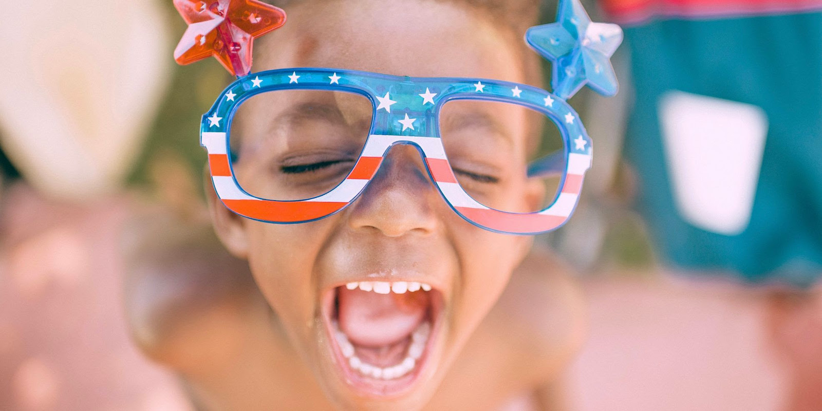 A young Black child wears American-flag themed glasses and smile-screams into the camera.
