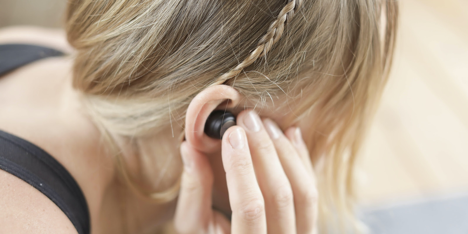A photograph of a young white woman with blond hair from behind shows her holding an earpiece into her ear.