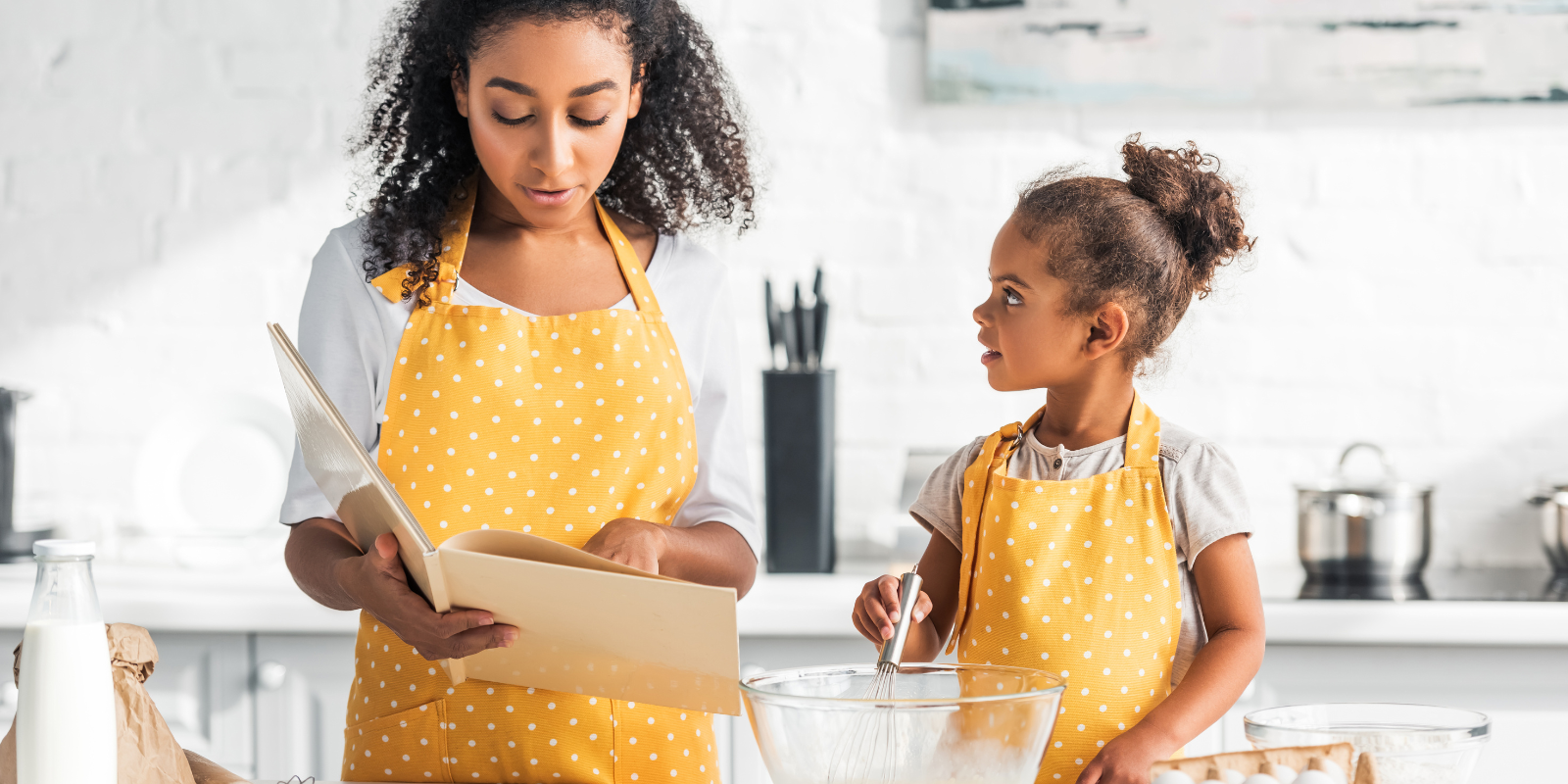 A young Black teenage girl with curly brown hair holding a cookbook and a small Black toddler with curly brown hair tied up both wearing yellow polka-dotted aprons stand at a kitchen counter covered with cooking tools.