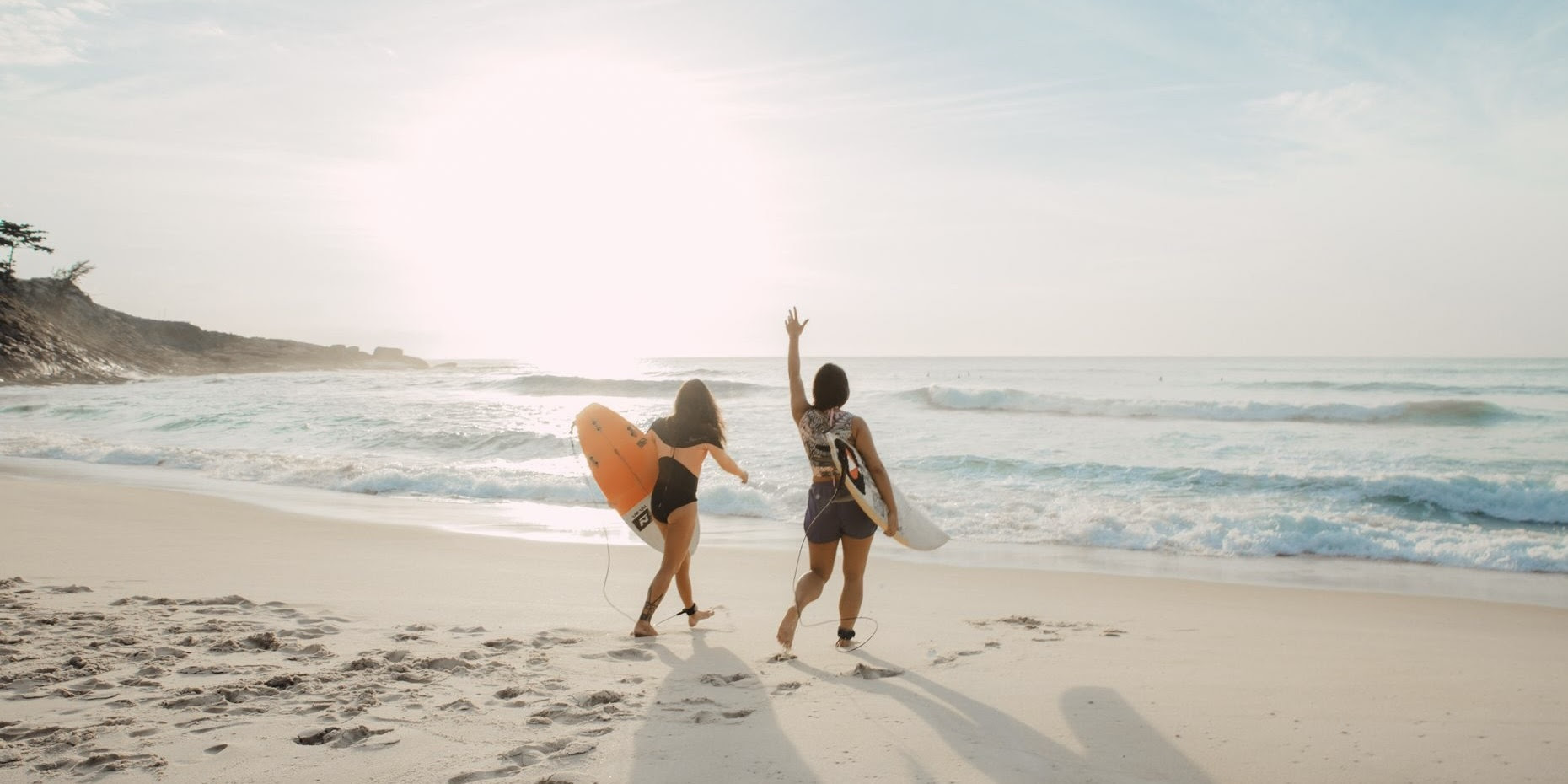 A photograph of a beach at sunrise, with two white woman holding surf boards walking to the water.