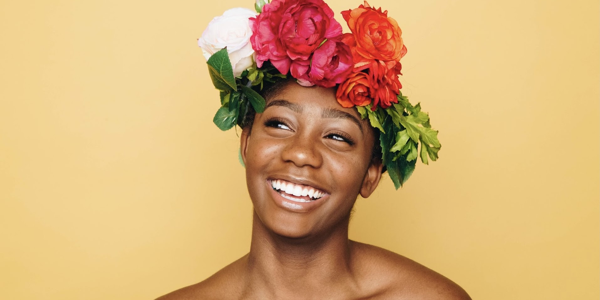 A photograph of a Black woman wearing a crown of big, bright flowers smiles and stands against a yellow wall.