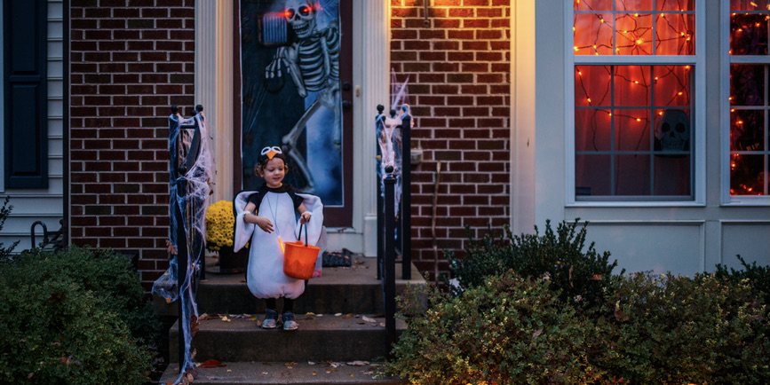 A color photograph of a small child dressed in a Halloween penguin costume with a plastic pumpkin basket leaves the doorstep of a house with a skeleton in the doorframe, its eyes lit up. In the windows of the house are orange lights.