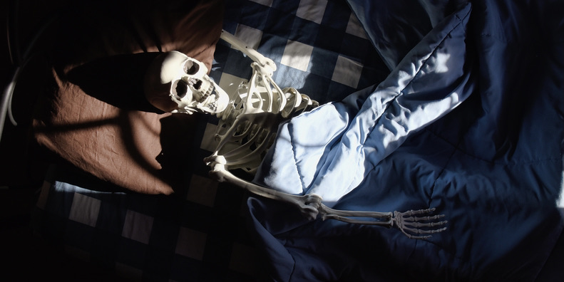 A color photograph of a (fake) skeleton lying in bed on blue checkered sheets under a blue comforter. 