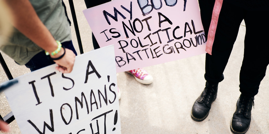 An overhead color photograph looking down on protest signs that read "My body is not a political battleground" and "It's a woman's choice" next to a person wearing black pants and boots.