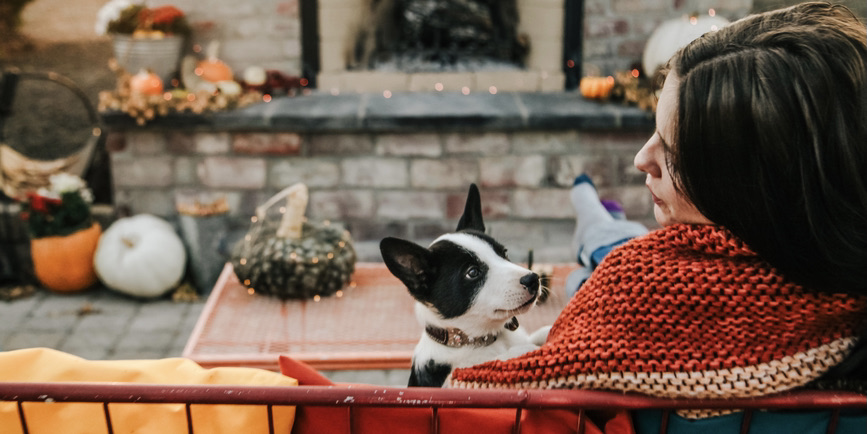 A color outdoor photograph of a white woman with brown hair wrapped in a colorful blanket looking into the face of a black-and-white puppy next to her while they sit in front of a pumpkin-adorned outdoor fireplace.