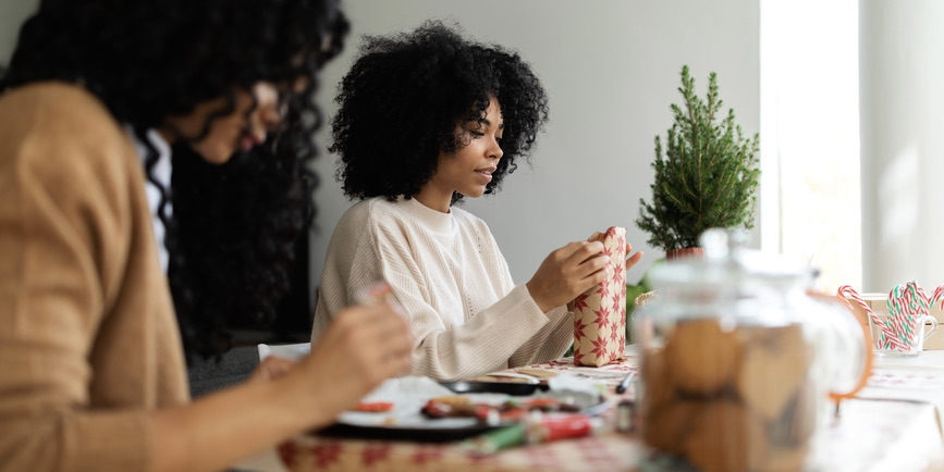 A color indoor photograph of two young Black women sitting at a kitchen table near a small evergreen tree, wrapping Christmas presents and frosting cookies.
