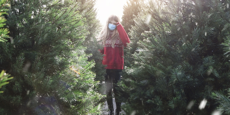 A color outdoor photograph of a young girl wearing jeans, a red sweater and medical face mask, standing between uncut Christmas trees.