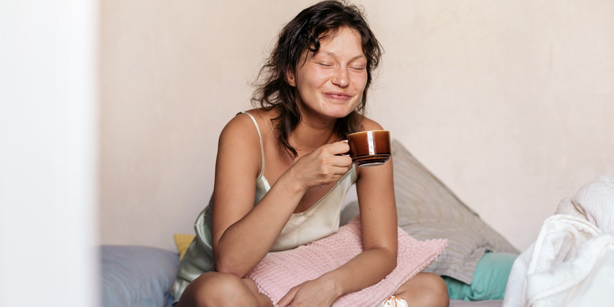 A color indoor photograph of a young white woman with short hair, sitting on a bed in a tank top with pillows around her, closing her eyes and smiling as she sips from a mug of tea.